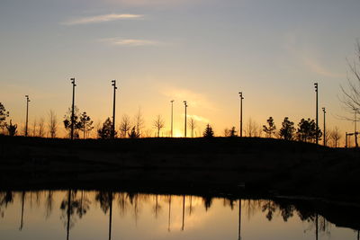 Silhouette factory by lake against sky during sunset