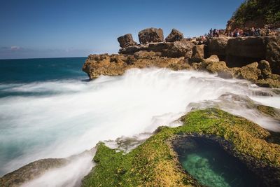 Waves splashing on rocks at beach against blue sky during sunny day