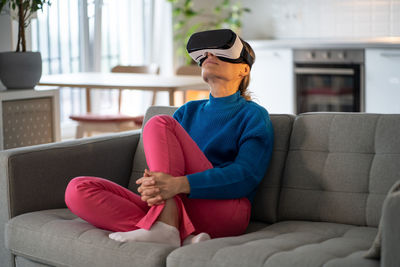 Intrigued european woman wearing virtual reality glasses relaxing after day at work sits on sofa