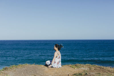Side view of woman sitting on cliff by beach against clear sky