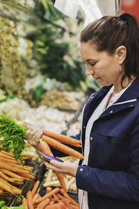 Side view of woman using smart phone while buying carrots in supermarket