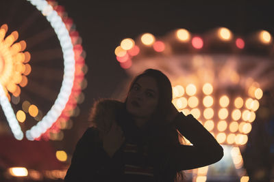 Portrait of woman standing at illuminated amusement park during night