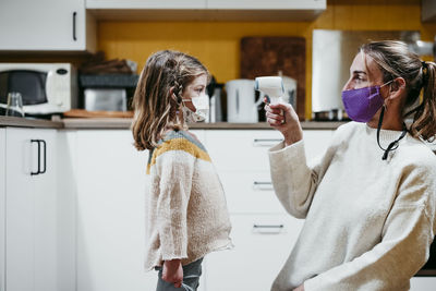 Mother wearing face mask checking temperature of daughter while sitting at home