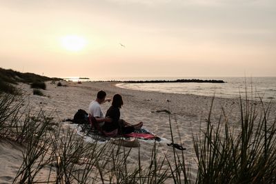 Couple sitting on beach against sky during sunset