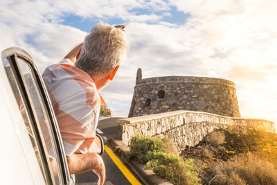 Rear view of senior man peeking out from car window against sky