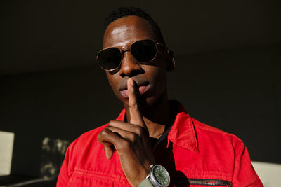 Portrait of young man in red jacket and sunglasses with finger on lips outdoors