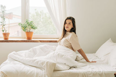 Young attractive brunette sits on the bed by the window after waking up in the morning.