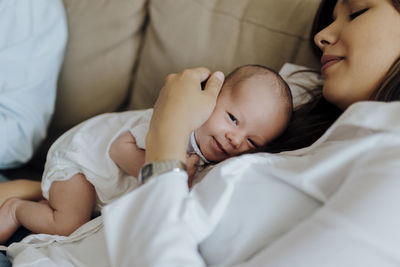 Happy newborn baby boy looking at the camera on top of his mother lying on the sofa