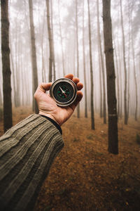 Close-up of hand holding navigational compass against bare trees in forest during foggy weather