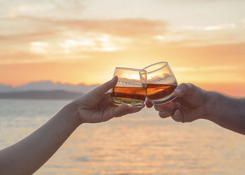 Cropped hands of couple toasting glasses at beach against orange sky