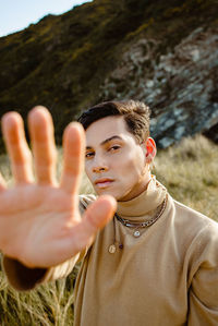 Young man in stylish outfit with fancy accessories standing on grass and looking at camera on sunny day in meadow