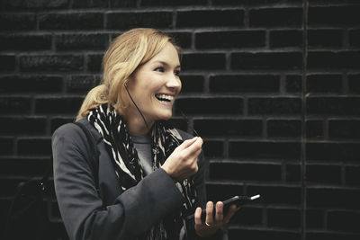 Smiling mid adult woman using hands-free device against brick wall