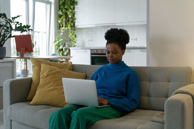 Young african american woman freelancer typing article in laptop sits on sofa in modern apartment