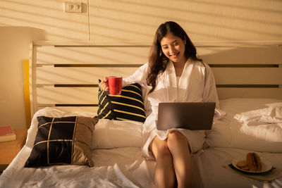 Young woman using phone while sitting on bed at home