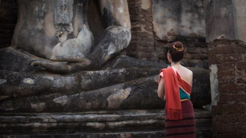 Rear view of woman wearing traditional clothing praying by statue at temple