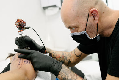 Crop male tattooist with machine making tattoo on arm of woman lying on table in salon