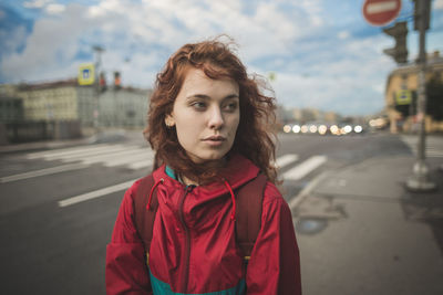 Serious millennial redhead female with curly hair wearing red jacket looking away while standing alone against road and historic buildings in windy day in saint petersburg in russia