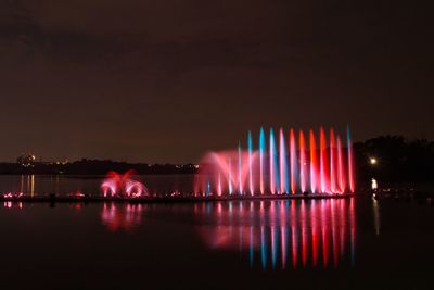 Illuminated fountain by lake against sky at night