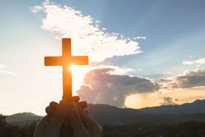 Cropped hand of person holding religious cross against sky during sunset