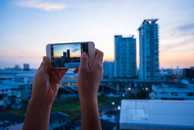 Cropped hands of woman photographing cityscape against sky during sunset
