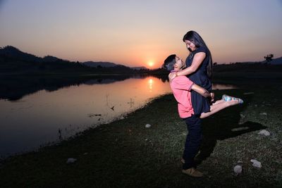 Full length of young man lifting woman while standing on grass field by lake against sky during sunset