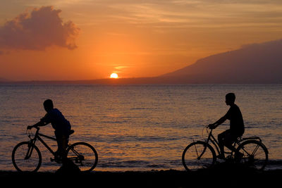 Silhouette boys cycling on shore at beach during sunset