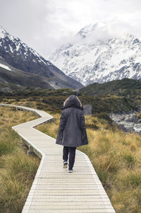 Rear view of woman wearing warm clothing while walking on boardwalk against snowcapped mountains