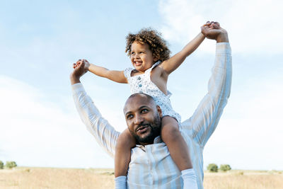Cheerful african american father with cute little daughter on shoulders playing in field in summer and having fun looking away