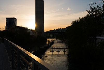 Silhouette bridge over river by buildings in town at sunset