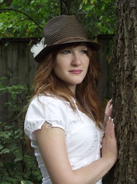 Young woman wearing hat standing by tree trunk in park