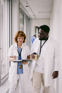 Senior female doctor discussing over medical record with young colleague while walking in hospital corridor