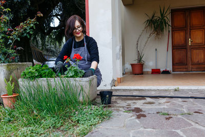 Woman in apron planting floers in the pot in the garden in front of the house