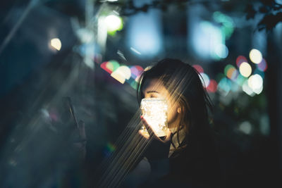 Woman using smart phone while holding glowing jar in city at night