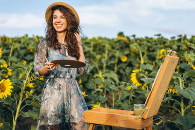 A young woman with curly hair and wearing a hat is painting in nature. 