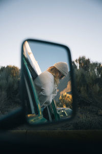 Young woman peeking through window reflecting on mirror against sky