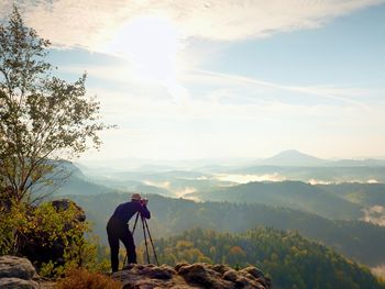 Nature photographer stay at tripod on summit and thinking. hilly foggy landscape 