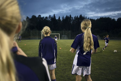 Rear view of girls standing on soccer field against sky at dusk