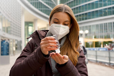 Woman wearing protective medical mask using alcohol gel sanitizing her hands outside shopping mall