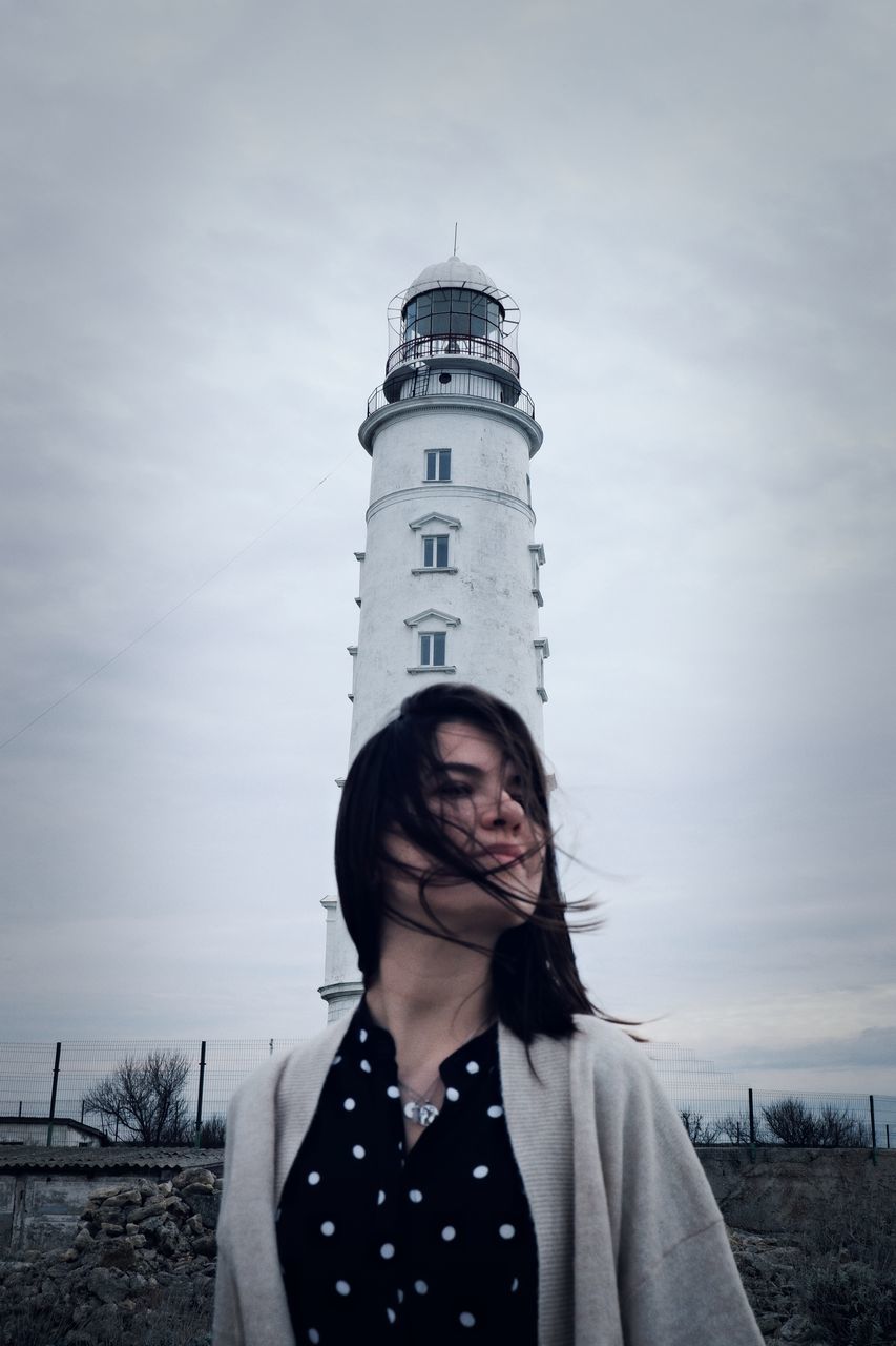 one person, sky, young adult, portrait, adult, standing, blue, clothing, lighthouse, women, nature, white, front view, cloud, architecture, person, human face, looking at camera, black, tower, waist up, winter, leisure activity, looking, long hair, emotion, smiling, casual clothing, hairstyle, day, outdoors, built structure, lifestyles, land, building exterior, spring, happiness, protection