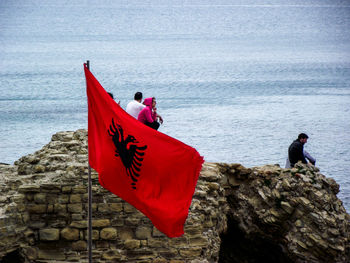 Red flag against people sitting on rock by sea