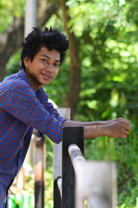 Portrait of young man leaning on railing against trees