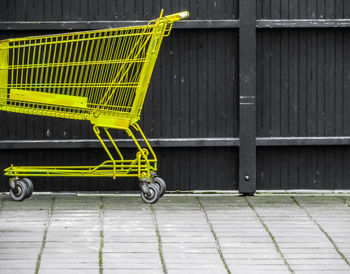 Empty yellow shopping cart on footpath against fence
