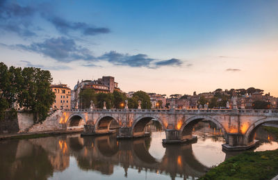 Arch bridge over river by buildings against sky at sunset