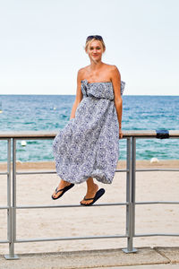 Portrait of woman sitting on railing by beach against sky