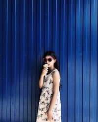 Portrait of girl eating ice cream while standing against blue corrugated iron