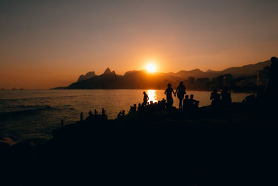 Silhouette people on shore at arpoador beach against orange sky