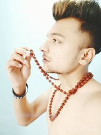Close-up of shirtless young man holding rudraksha necklace while looking away against white wall