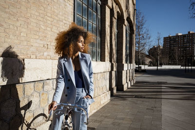 Young woman in blazer standing with bicycle by building during sunny day