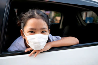 The little asian girl wears a hygiene mask and pokes her head out of the car window.