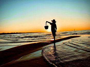 Silhouette woman holding bucket while standing at beach against sky during sunset